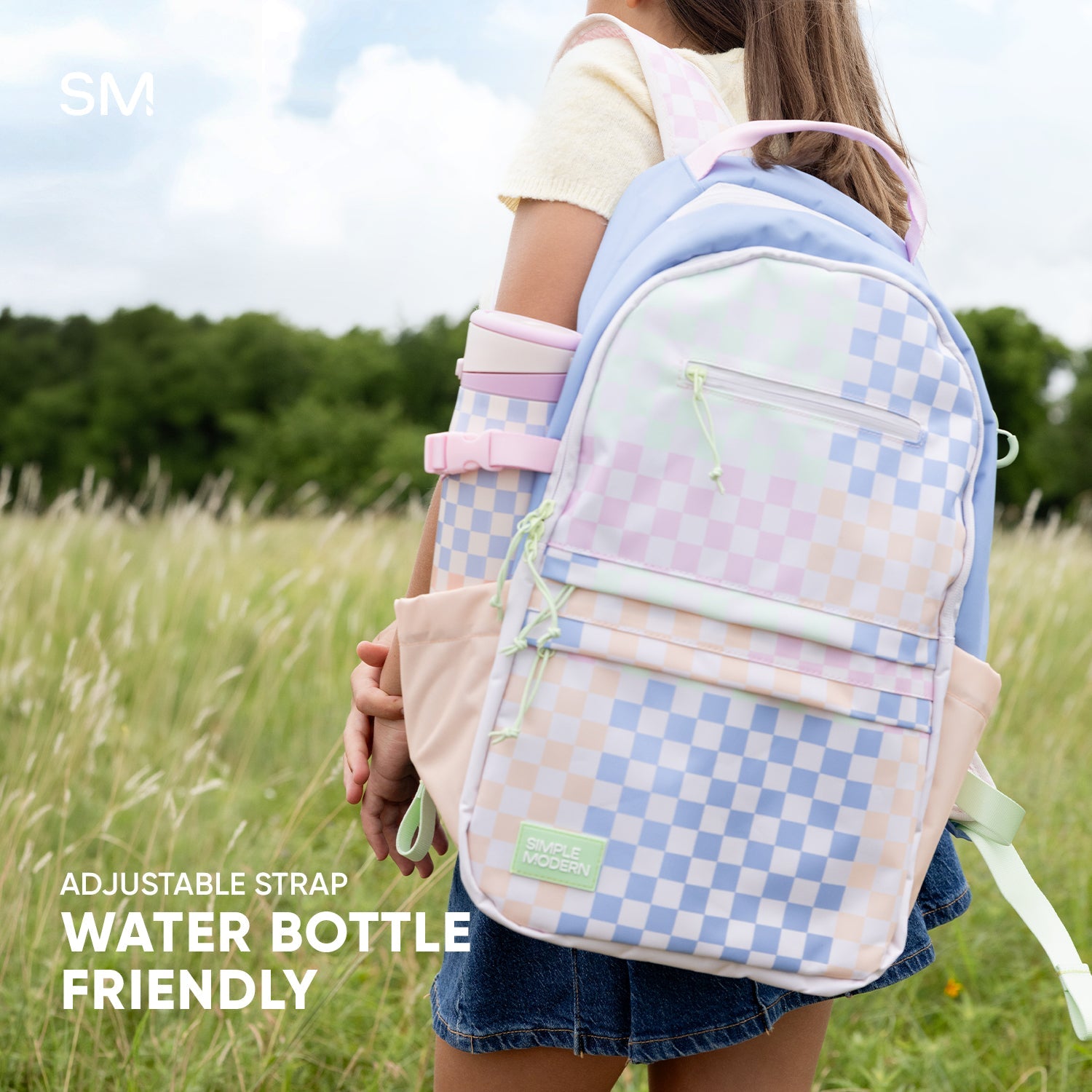 Young person wearing a Simple Modern 25L pastel checkerboard backpack with pink straps and lime green accents in an outdoor field setting with trees and blue sky.