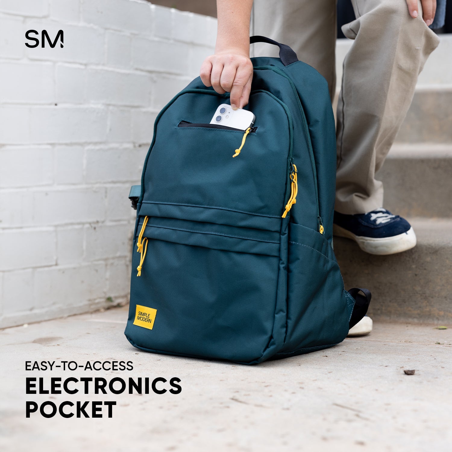 Person holding teal SM backpack with golden yellow accents against white brick wall, displaying smartphone in easy-access top pocket, wearing tan top and navy sneakers.