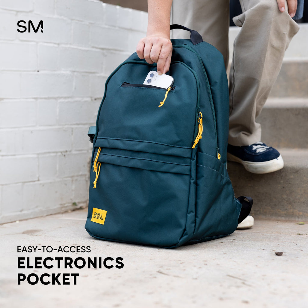 Person holding teal SM backpack with golden yellow accents against white brick wall, displaying smartphone in easy-access top pocket, wearing tan top and navy sneakers.